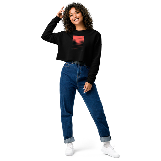 A person with curly hair is smiling and posing against a black background while wearing a Layers Supply WS001 ▪ Crop Sweatshirt with a red gradient design, blue high-waisted jeans, and white sneakers. One hand is touching their trendy cut, and the other is relaxed by their side.